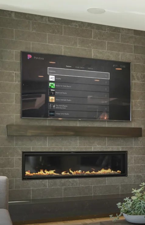 A living room TV above a fireplace showing a music service